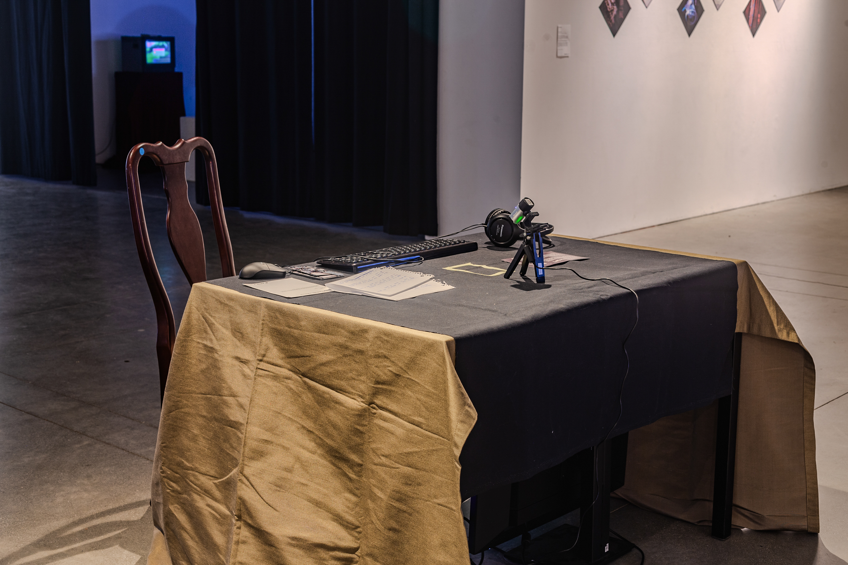 A table sits in the middle of a white space, there is table cloth on it with playing cards and an instruction manual. There is a webcam connected to a 6 inch tripod pointing towards the cards with headphones. Around the back of the table is an upholstered chair.